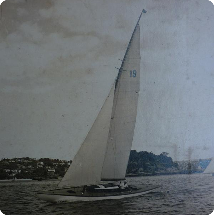 A vintage photo of a Classic Sailing Yacht with the number 19 on its tall sail gliding on the water, a coastal town and trees in the background under a cloudy sky.