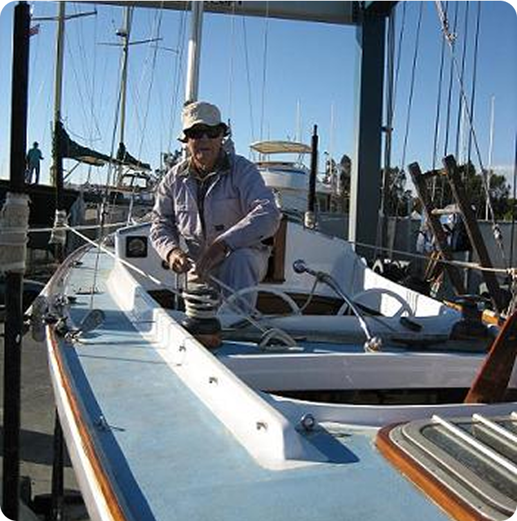A person wearing sunglasses, a hat, and light-coloured clothing is adjusting equipment on the deck of a docked Classic Sailing Yacht on a sunny day. Sailboat masts and a marina are visible in the background.