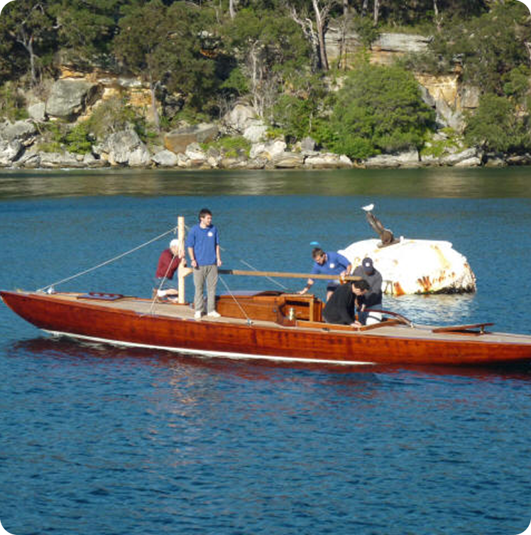 A group of people stand and sit on a polished wooden Schärenkreuzer, a classic sailing yacht, floating on calm blue water, with a rocky, tree-lined shore and a large white rock visible in the background.