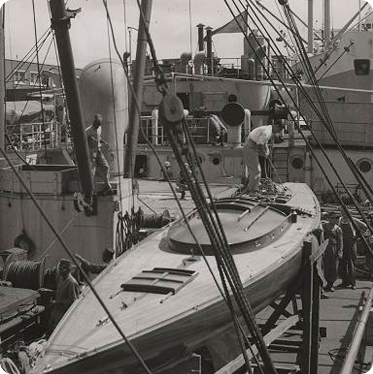 Black and white photo of workers preparing a Schärenkreuzer wooden boat on the deck of a larger ship at a dock, with ropes and equipment visible, capturing the timeless craft of Classic Sailing Yachts.