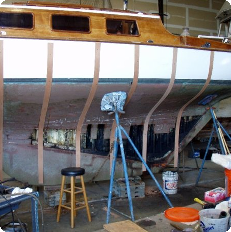 A wooden Square Metre Yacht in a garage is under repair, with new wooden ribs attached to its hull. Nearby are tools, paint tins, and a cloth-covered stool, while the unfinished hull reveals the classic yacht's underlying structure.