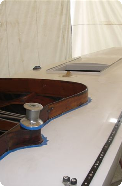 A close-up view of a Classic Sailing Yacht's deck inside a marquee, showing a wooden structure, a metal winch, a hatch, and some fittings on a white surface. The background is a fabric enclosure.
