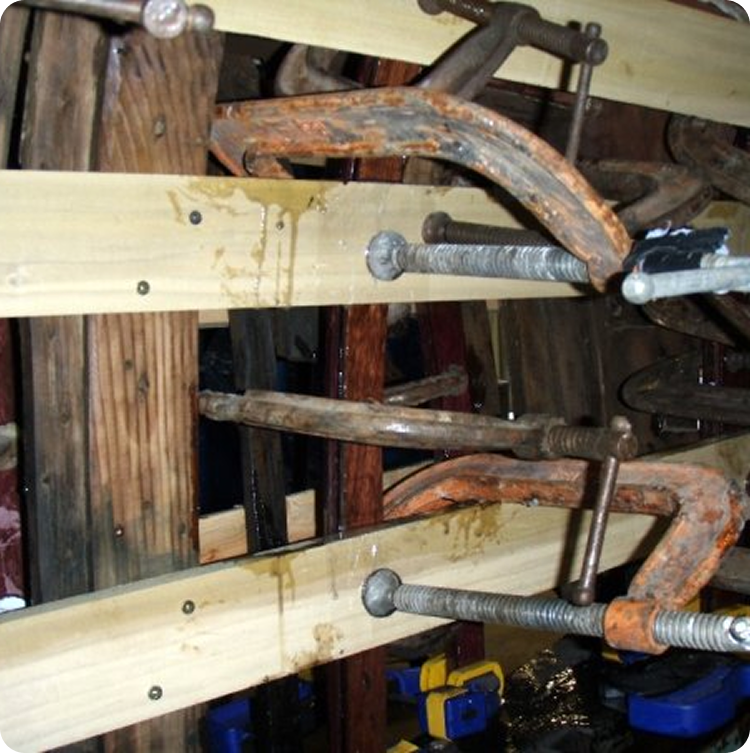 Several large metal cramps hold wooden boards together, water droplets visible on the wood and cramps, suggesting a wet environment or glue drying—likely part of restoring Classic Sailing Yachts or maintaining elegant Square Metre Yachts.