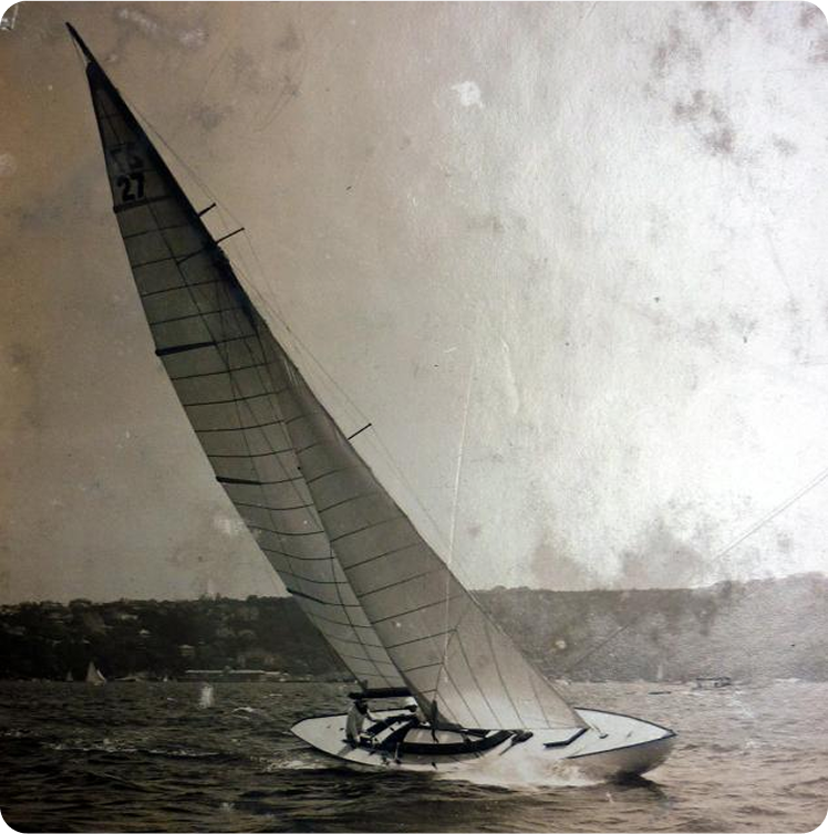A vintage photograph of a single Classic Sailing Yacht with a tall mast gliding on the water at an angle, with a hilly shoreline and faint buildings visible in the background.