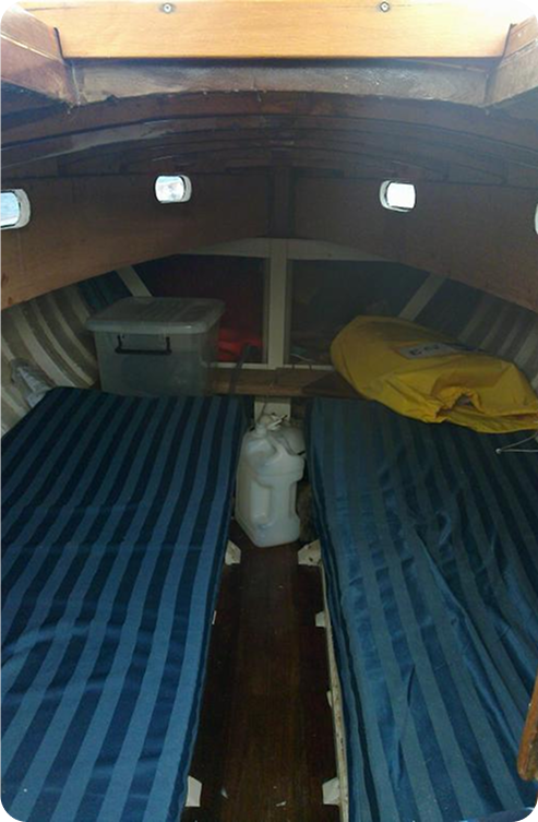 The image shows the interior of a Schärenkreuzer boat cabin with two narrow blue-striped beds on either side, a yellow bag and plastic storage box at the end, and a white container on the dark wooden floor between the beds.