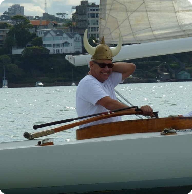 A man wearing a horned Viking helmet and sunglasses smiles while sailing a Skerry Cruiser on a body of water, with buildings and trees visible in the background.