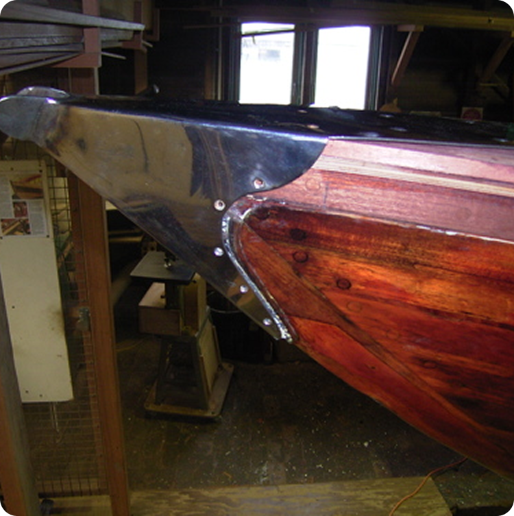 The photo shows the bow of a wooden boat, resembling classic Skerry Cruisers, with shiny metal plating secured with bolts at the tip. The boat is inside a workshop with tools and equipment visible in the background.