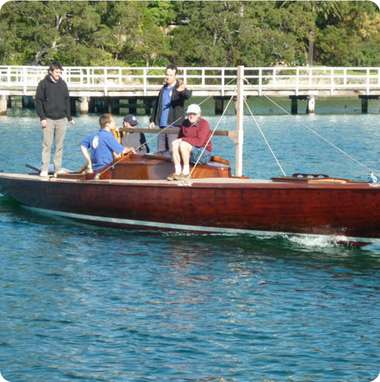Five people are riding a wooden boat reminiscent of Classic Sailing Yachts on calm, blue water near a pier with trees in the background. Some are seated whilst others stand and talk, enjoying the sunny day.