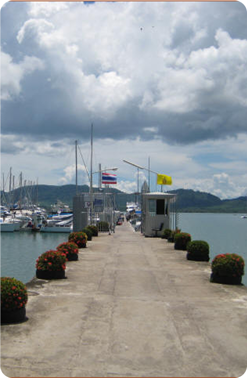 A concrete pier lined with potted plants extends into the water, with classic sailing yachts and moored sailboats on the left, two flags flying at the end, and cloudy skies above.