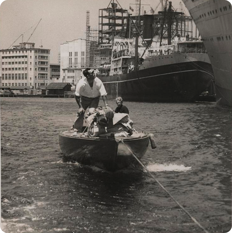 A small boat with two people is being towed in a harbour, passing by classic sailing yachts and large docked ships, with industrial buildings and cranes in the background. The scene appears to be from a past era.