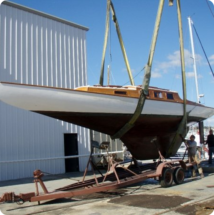 A classic sailing yacht is being lifted by straps attached to a crane, above a trailer at a marina, with a white building and blue sky in the background.