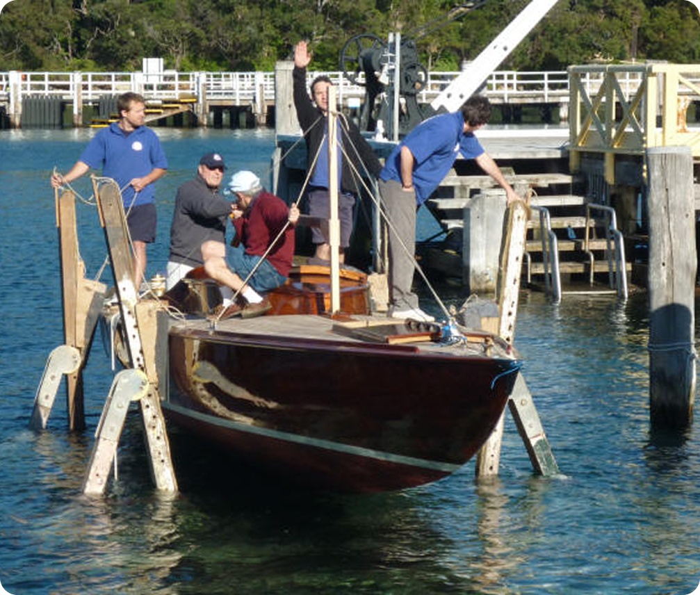 Five people are working together to launch or retrieve a small wooden Skerry Cruiser at a quay. One person waves whilst others work on or around the boat, which is being held by a lifting structure in the water.