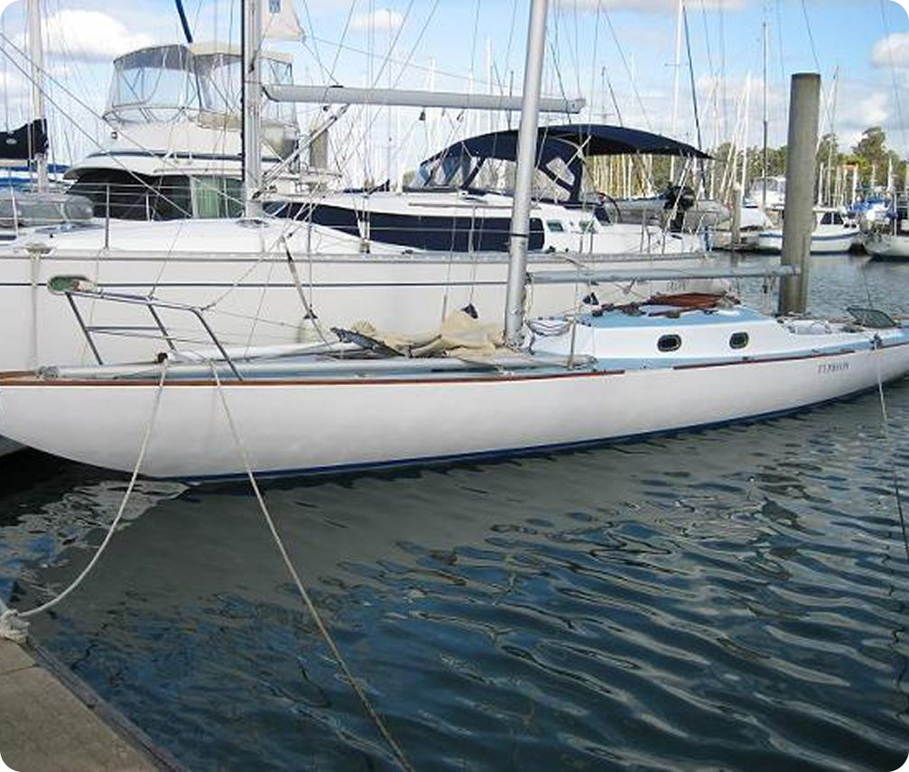 A small white Schärenkreuzer is moored in a marina, tied to the pier with ropes. Other larger boats and yachts are visible in the background on a calm, sunny day.