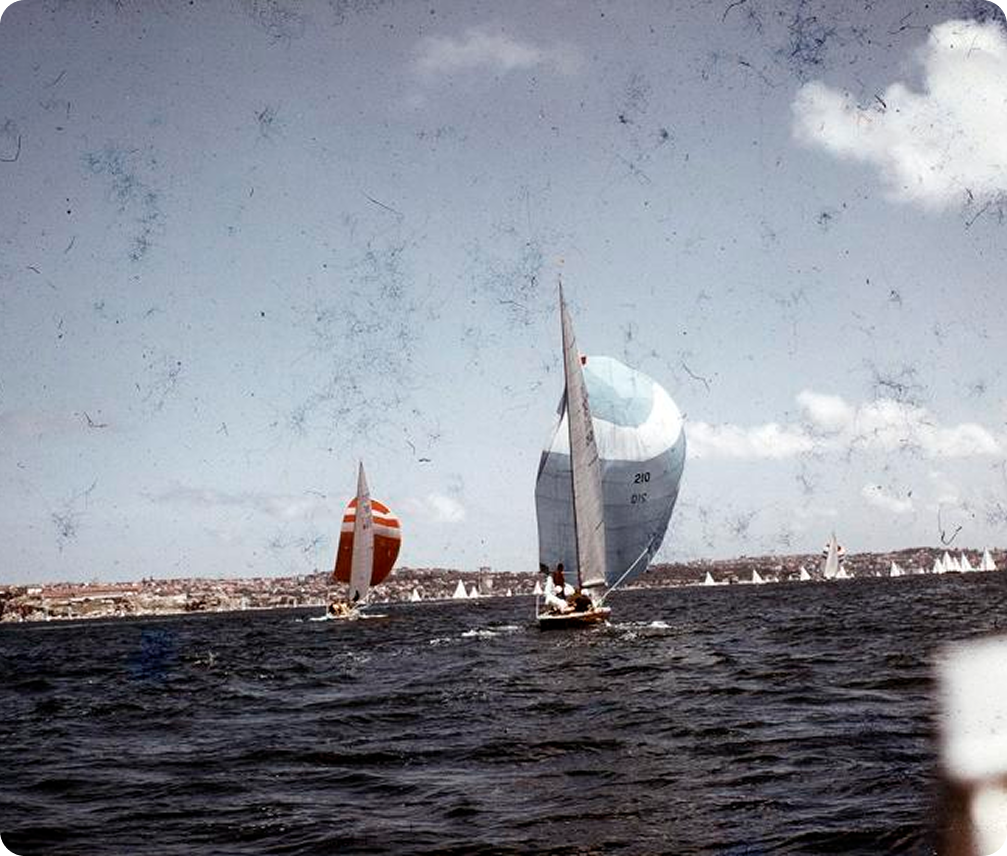 Sailing boats, including elegant Square Metre Yachts with colourful sails, glide across a dark blue body of water under a partly cloudy sky, with a distant shoreline and additional sailing boats visible in the background.