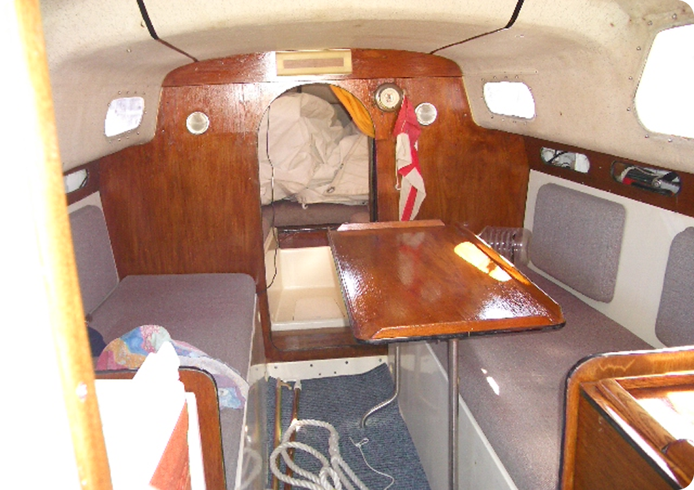 The image shows the cosy interior of a Schärenkreuzer sailboat cabin with wooden walls, a folding table, cushioned benches, coiled rope on the floor, and a berth with white sails stored at the back. Sunlight shines on the table.