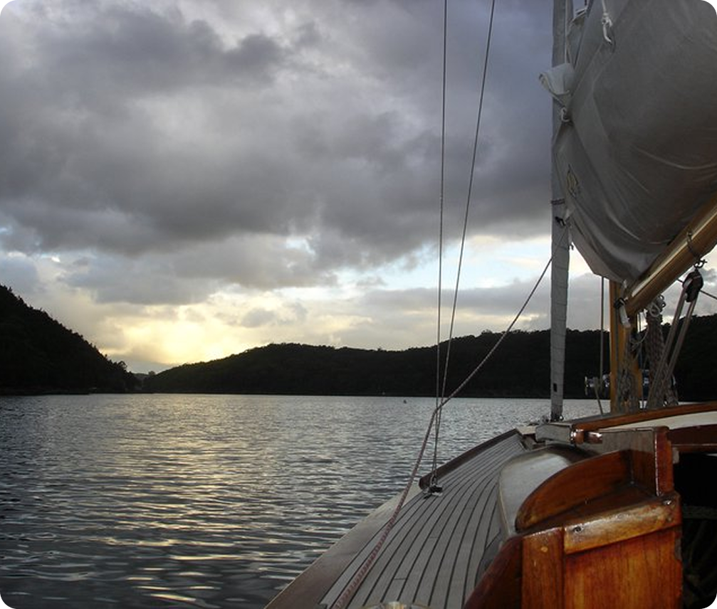 A view from a Classic Sailing Yacht on a calm lake at sunset, with dark hills in the distance and dramatic clouds overhead. The deck and part of the sail are visible in the foreground, capturing timeless elegance on the water.