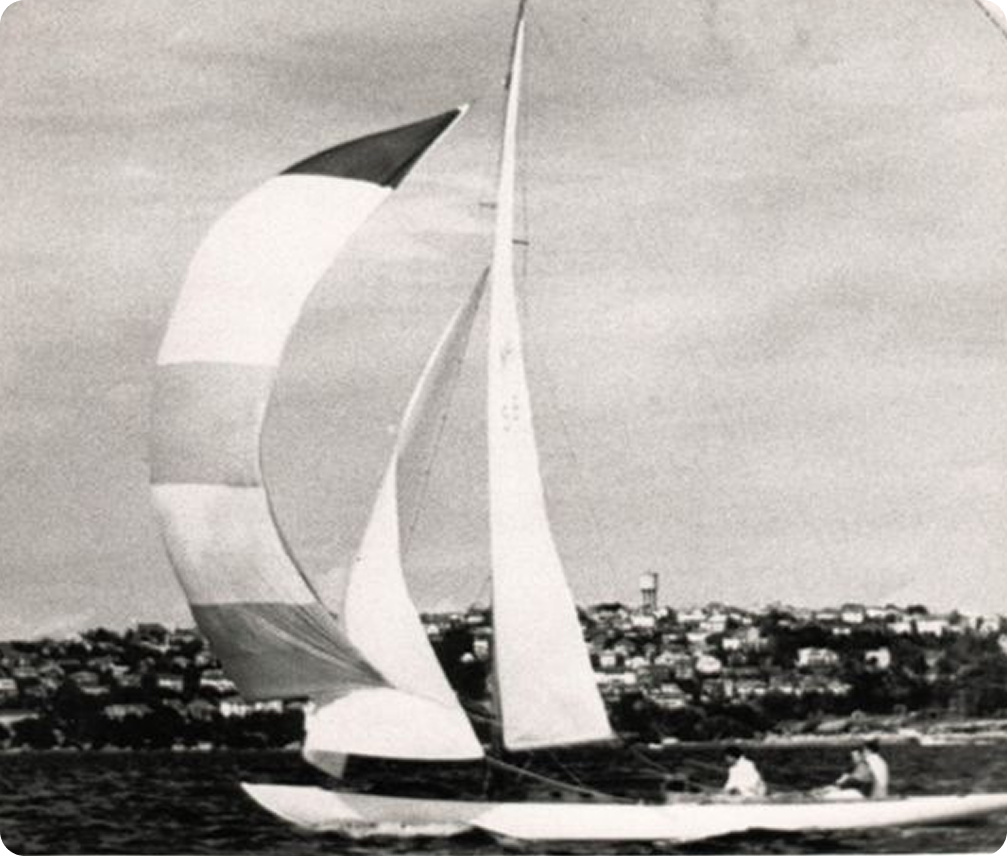 A black and white photo of a classic sailing yacht with large, billowing sails on the water. Two people are on board, and a town with many buildings is visible on the shoreline in the background.