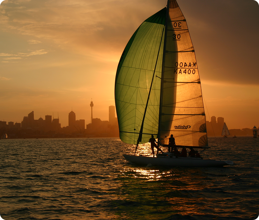 A classic Square Metre Yacht with green sails glides across the water at sunset, silhouetted against a city skyline. The golden sky and shimmering reflections capture the timeless elegance of these renowned vessels.