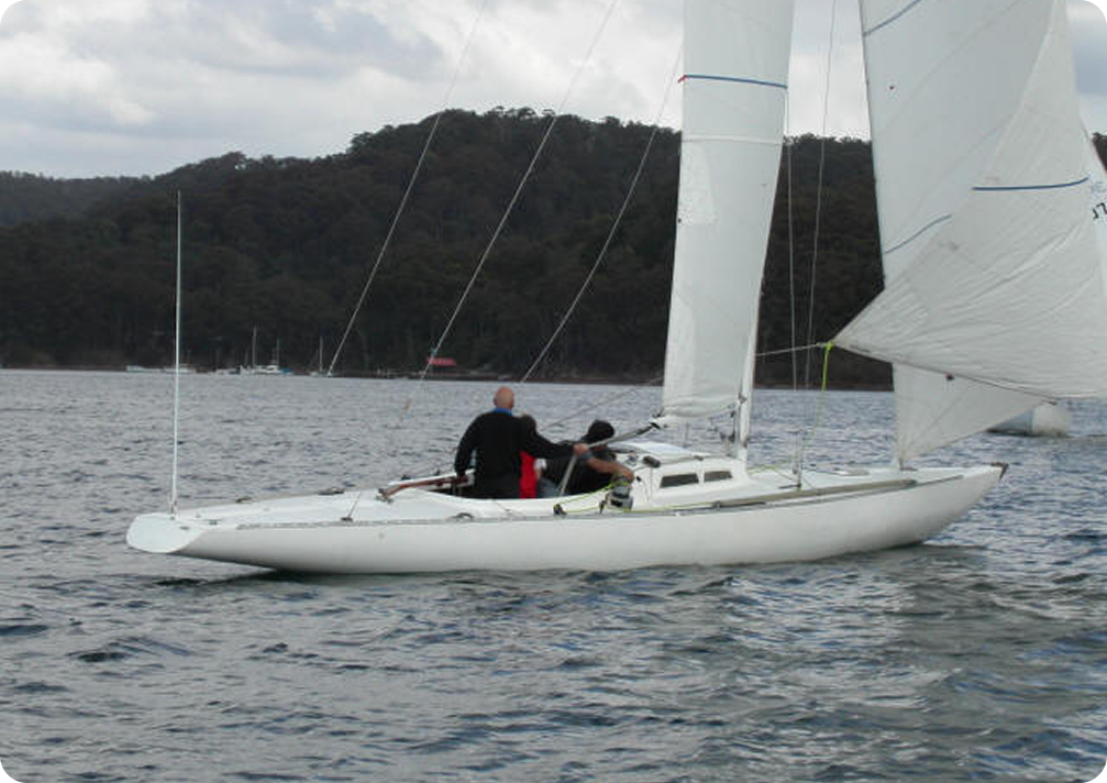 Two people are sailing a white Schärenkreuzer on calm water, with a forested shoreline and other classic sailing yachts visible in the background under a cloudy sky.