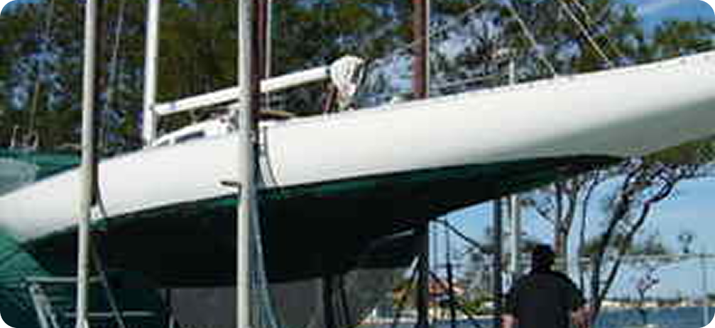 A person stands near a white Skerry Cruiser sailboat propped up on supports, with trees and a blue sky in the background.