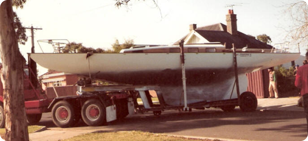 A large Classic Sailing Yacht is being transported on a flatbed lorry through a residential neighbourhood, with houses, trees, and a person standing nearby.