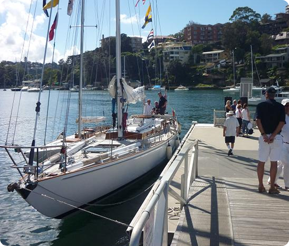 A white Skerry Cruiser sailboat moored at a marina with people standing on the pier and nearby; houses and trees are visible in the background under a partly cloudy sky.
