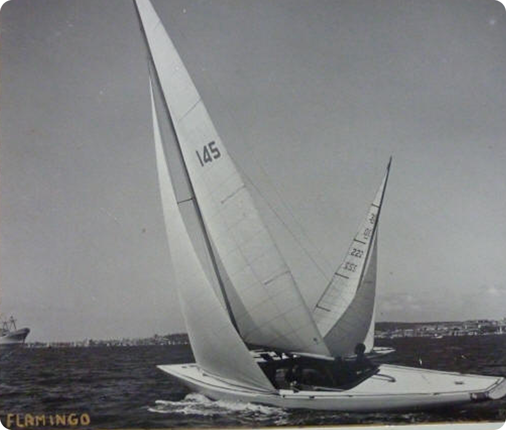 A vintage black-and-white photo of a Square Metre Yacht marked 145 gliding on the water, with several people on board. The word FLAMINGO appears at the bottom left, and city buildings are visible on the distant shore.