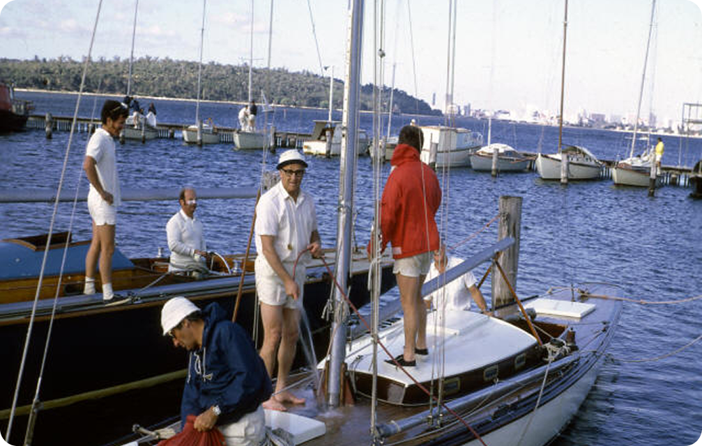 Four men stand on and around a moored Schärenkreuzer sailboat at a marina. One wears a navy jacket and white cap, another a red hoodie, and two wear white shirts and shorts. Other boats and a distant shoreline are visible.