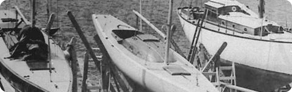 Black and white photo of three moored boats, featuring a sleek Skerry Cruiser in the centre, surrounded by two larger Classic Sailing Yachts on calm water.