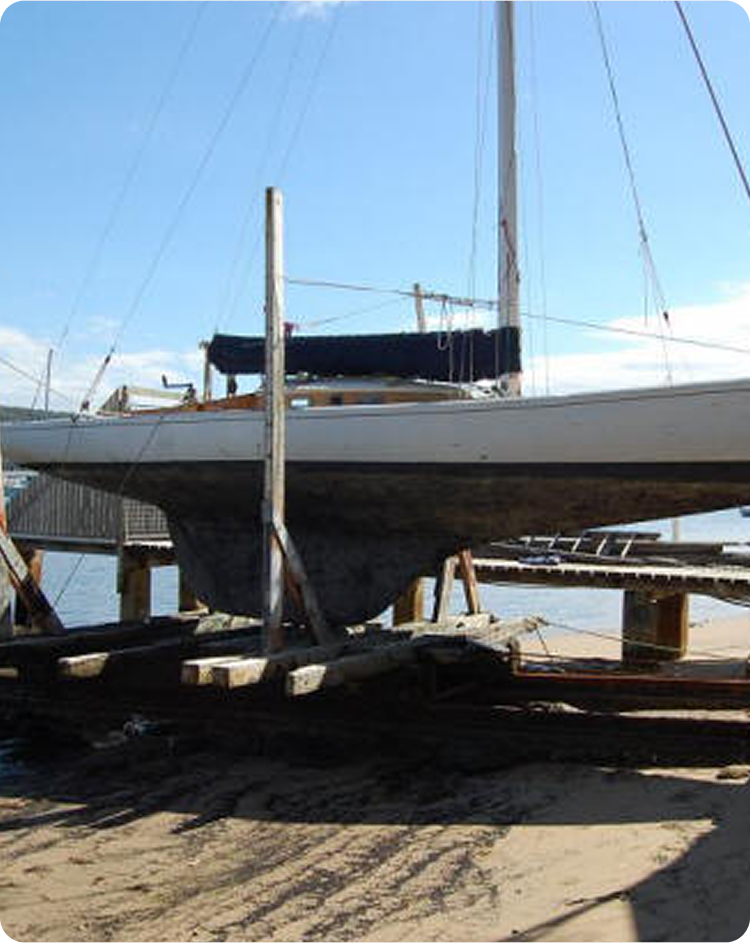 A sailboat, reminiscent of Classic Sailing Yachts, is propped up on wooden supports at a sandy boatyard near the water, with a clear sky and jetties visible in the background.