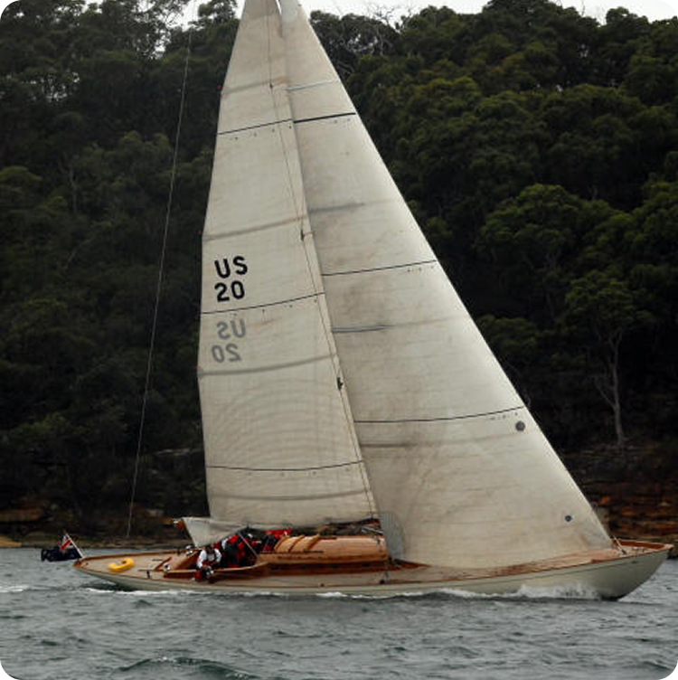 A classic wooden Skerry Cruiser with the sail marked US 20 glides on the water near a forested shoreline, with a few people on board and an overcast sky above.