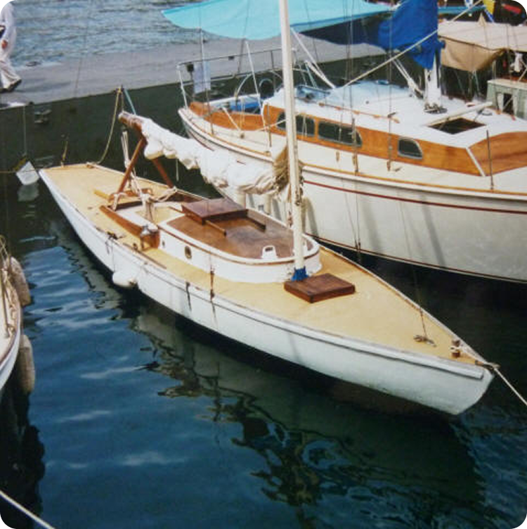 A small white sailboat with a wooden deck, evoking classic sailing yachts, is moored next to a larger cabin cruiser. Blue tarpaulins provide shade as the mast and rigging stand tall, reflected beautifully in the water.