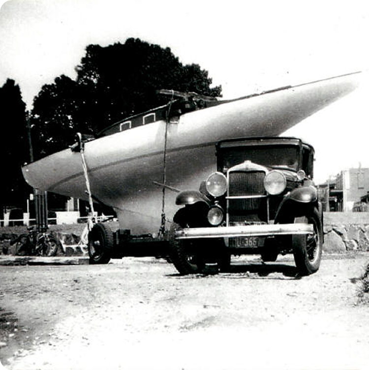 A vintage black-and-white photo shows an old car towing a large Square Metre Yacht on a trailer, parked on a dirt surface with trees and buildings in the background.