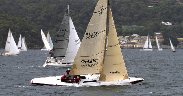 Sailing boats, including elegant Classic Sailing Yachts with tan and white sails, race across a choppy stretch of water, lush green hills and a few buildings in the background. Several people are on board the nearest sailing boat.