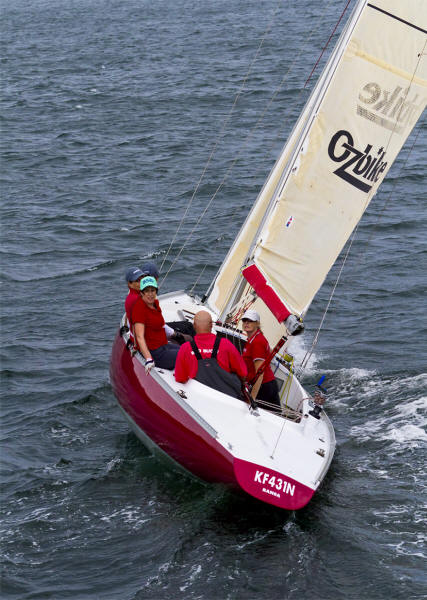 Four people in red shirts sail a classic sailing yacht—a small red and white boat with a cream sail—on open water. The sail reads Gzbike, and the registration KF431N is on the hull, evoking the charm of square metre yachts.