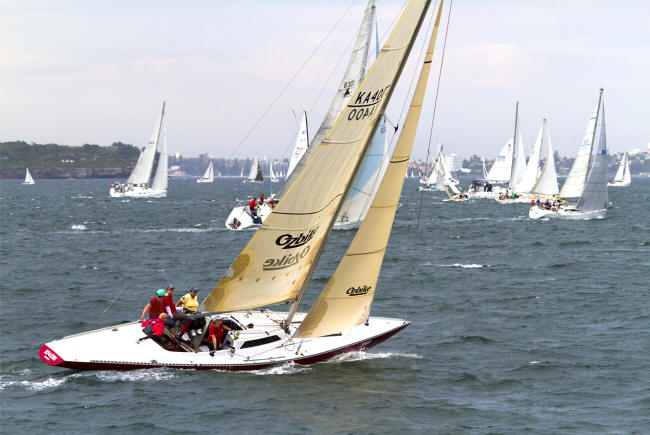 A sailing boat with a group of people in life jackets glides across choppy water, joined by other Square Metre Yachts in the background under a cloudy sky.