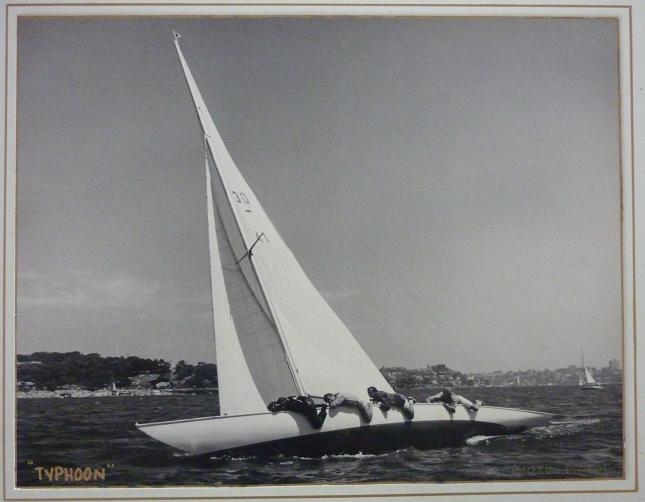 A vintage black-and-white photo of the sailboat Typhoon, a classic Square Metre Yacht, leaning dramatically on the water as crew members hang over the side to balance it. A distant shoreline and another sailboat are visible in the background.