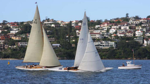 Two Square Metre Yachts with large white sails race on a calm body of water near a shoreline lined with houses, whilst a small motorboat floats nearby. Trees and hillside homes are visible in the background.