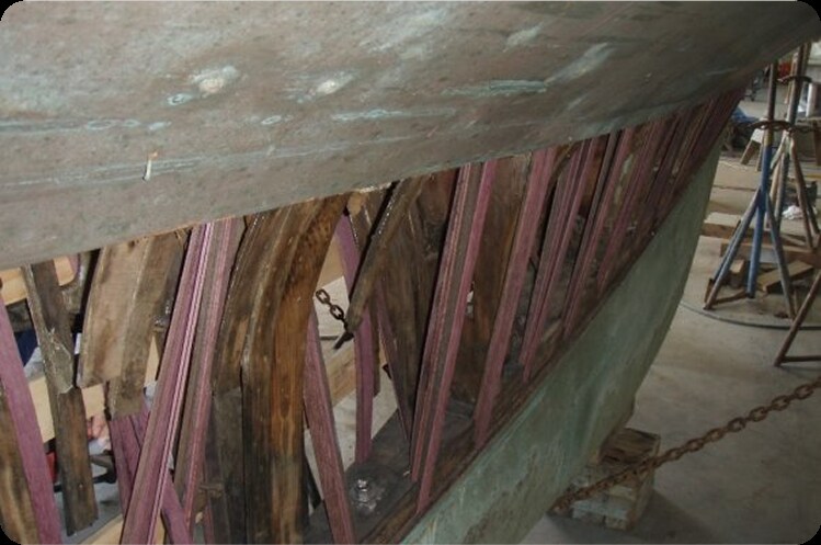 A close-up view of the underside of a Square Metre Yachts hull undergoing repair, showing exposed wooden ribs and beams with structural damage and missing planks. Chains and workshop tools are visible in the background.
