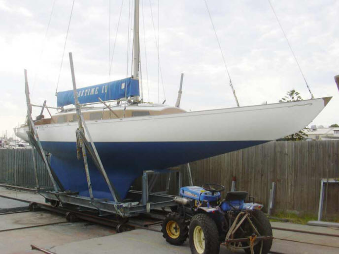A blue and white classic sailing yacht rests on a cradle near a wooden fence, with a blue tractor parked in front. The sky is overcast.