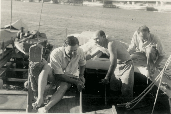 Three men on a wooden boat by a quay—one with legs crossed, another bare-chested and smiling, the third bending over. Classic Sailing Yachts and water with other boats are visible in the background.