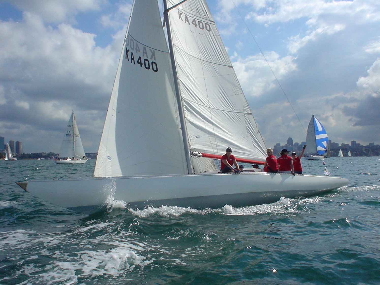 A white Square Metre Yacht with a crew of four in red shirts sails on choppy blue water. Other Skerry Cruisers are visible in the background beneath a partly cloudy sky.