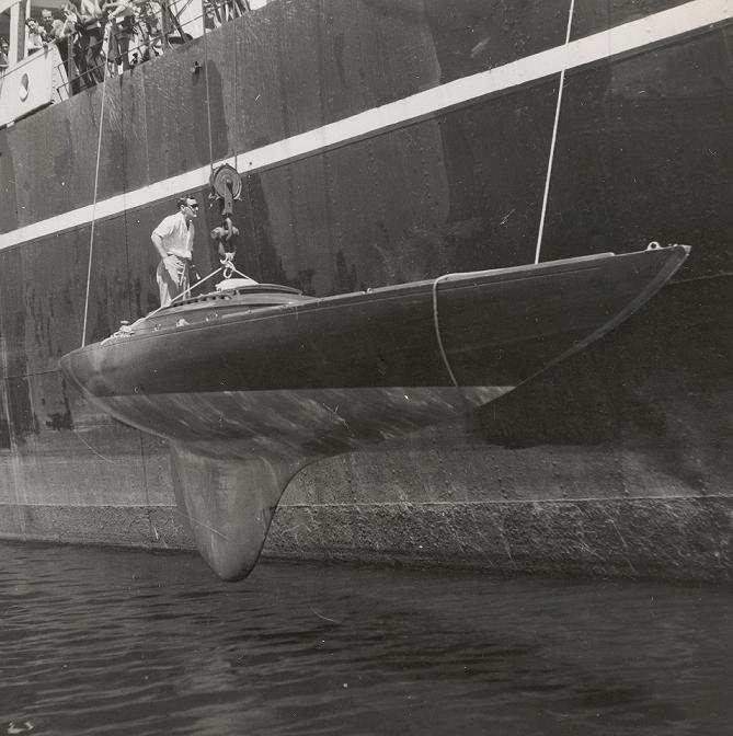 A man stands on a sleek Schärenkreuzer racing sailing boat being hoisted by a crane beside a large ship, with the classic sailing yacht partially above the water and ropes securing it from above.