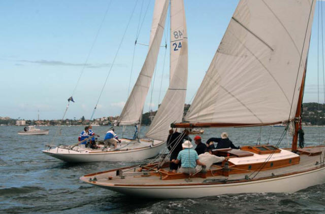 Two Schärenkreuzer sailboats race side by side on the water, each with several people on board. The background features a distant shoreline with buildings and trees beneath a blue sky.