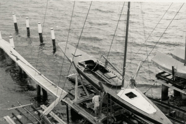 A black and white photo shows a Schärenkreuzer sailboat on a wooden dock hoist above the water, with a person standing nearby. Poles and another quay are visible in the background over the rippling water.