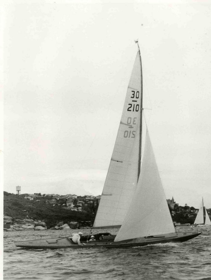 A classic sailing yacht with two people aboard glides on the water near a hilly shoreline dotted with houses and trees. The sailboat’s large sail is marked 30 210, resembling elegant Square Metre Yachts. Another boat sails in the background.