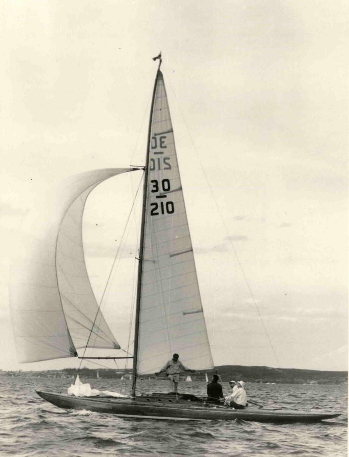 Black and white photo of a Schärenkreuzer, a classic sailing yacht, with three people aboard. Tall sails display “30” and “210” as they glide on the water, shoreline visible under a cloudy sky.