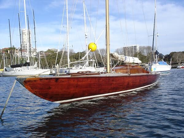 A classic wooden sailboat, reminiscent of Schärenkreuzer Skerry Cruisers, with a polished brown hull floats on calm water in a marina, surrounded by other sailboats and city buildings under a partly cloudy sky.