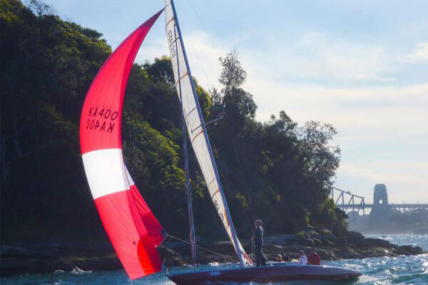 A small Schärenkreuzer with a red and white spinnaker sails near a wooded shoreline on a sunny day, several people on board and a bridge visible in the background.