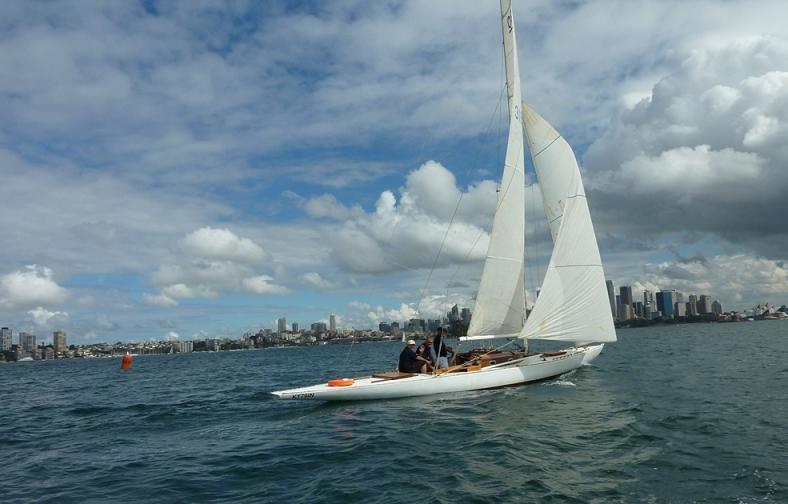 A white Schärenkreuzer sailboat with people on board glides across the water under a partly cloudy sky, with a city skyline visible in the background.